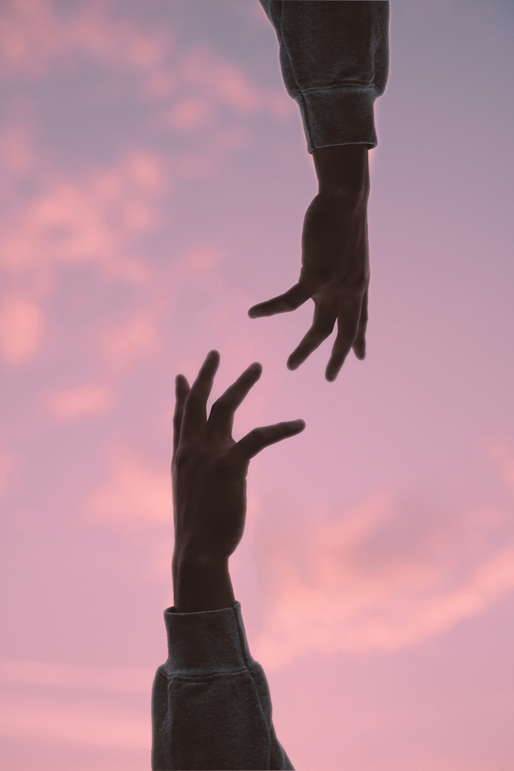 Two hands reaching toward each other against a pink sunset sky